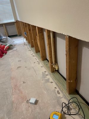 Room with lower drywall removed, exposed wall studs, cords, and mitigation equipment visible on the floor.