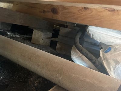 Crawlspace beneath a home showing wood framing, concrete block supports, plastic sheeting, and a flexible line or hose.