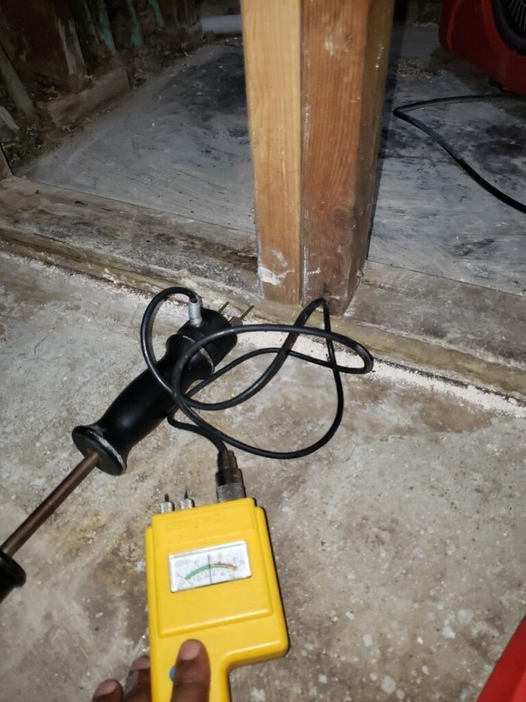 Handheld moisture meter testing exposed framing and concrete in a residential remediation area