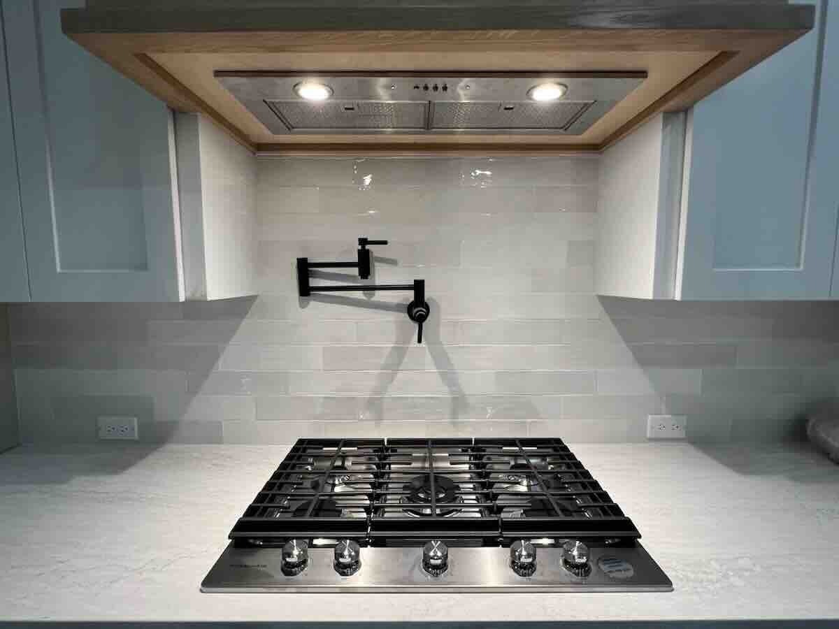 A modern kitchen stove with five burners, a stainless steel range hood with lights, and a black pot filler faucet mounted on a white tile backsplash. White countertops and cabinets surround the stove.