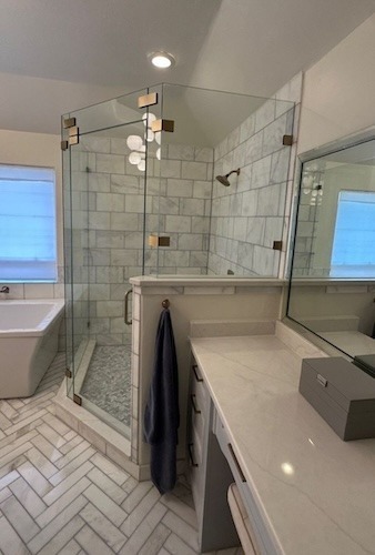 Modern bathroom with a glass-enclosed shower, white tile walls, a soaking tub, herringbone-patterned floor, large mirror, and counter with a gray box. A dark towel hangs by the shower door.